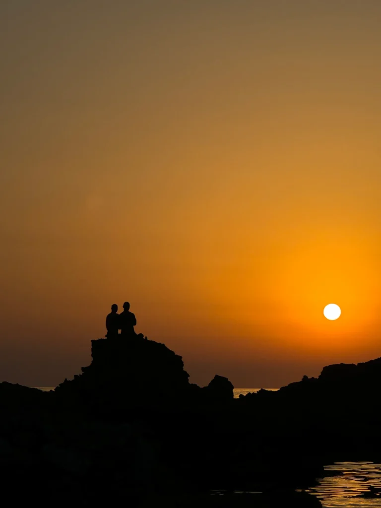 Silhouette of two people sitting on a rock along the coastline overlooking a deep orange sunset.
