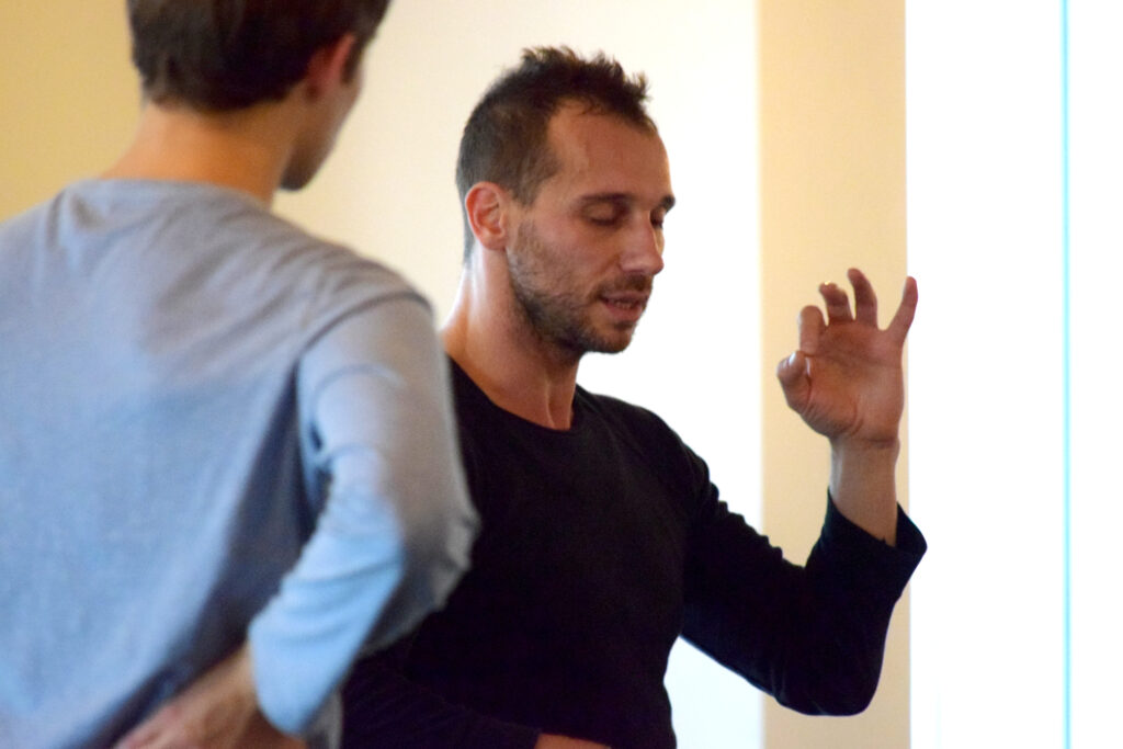 Man with eyes closed demonstrating technique to a young man in grey sweatshirt with his back to the camera.